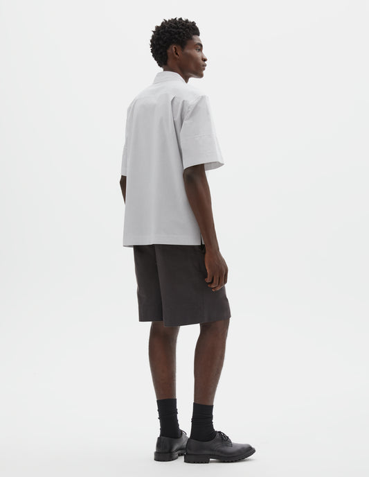 Full body image, back view of model wearing white shirt with grey stripes, panels on short sleeves with contrasting band at cuff and side split vents. Styled with dark brown shorts, black socks and black leather shoes.