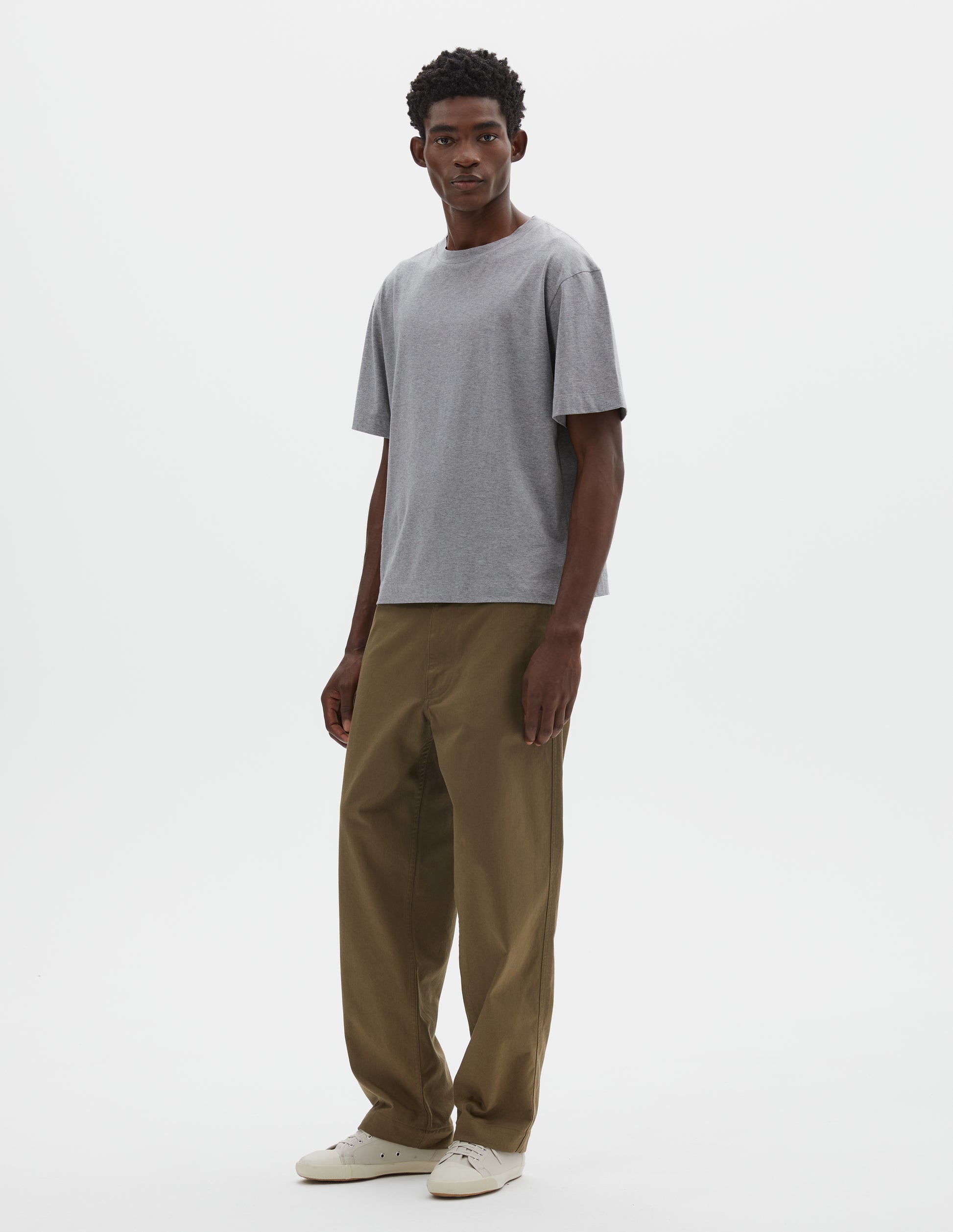 Full body image, front view of model wearing black cotton short sleeve t-shirt. Styled with green trousers and beige canvas trainers.