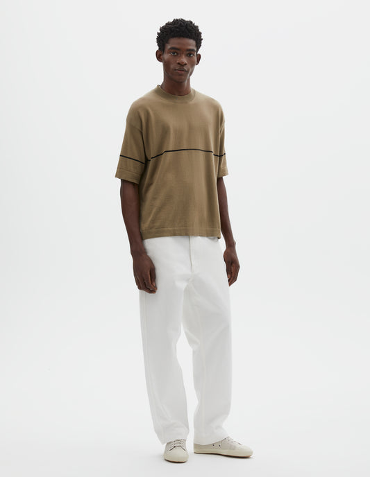 Full body image, front view of model wearing regular, generous fit, dark beige knitted t-shirt with deep rib crew neck, set in sleeves and single black stripe across body. Styled with white trousers and beige canvas trainers.