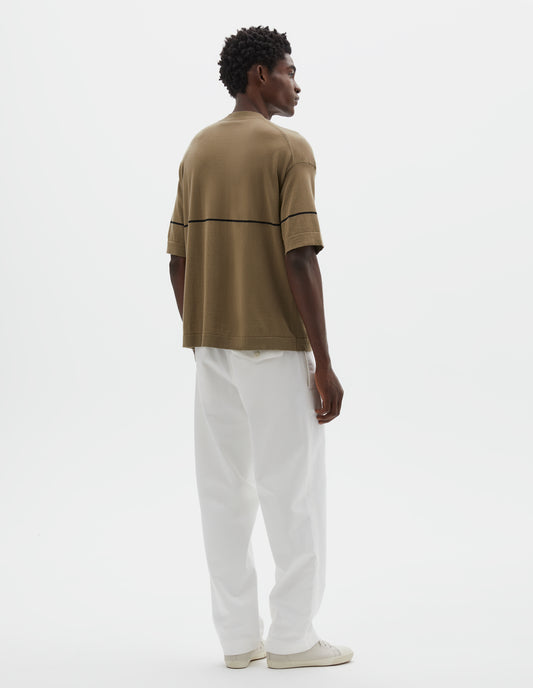 Full body image, back view of model wearing regular, generous fit, dark beige knitted t-shirt with deep rib crew neck, set in sleeves and single black stripe across body. Styled with white trousers and beige canvas trainers.