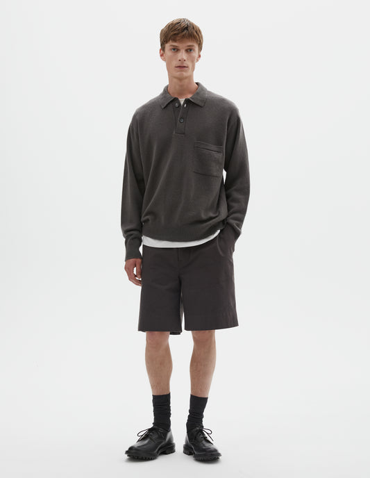 Full body image, front view of model wearing dark grey merino cashmere polo shirt with dropped sleeve, integral chest pocket, horn button closure and rib collar, cuffs and hem. Styled over white t-shirt, with brown shorts, black socks and black leather brogues.