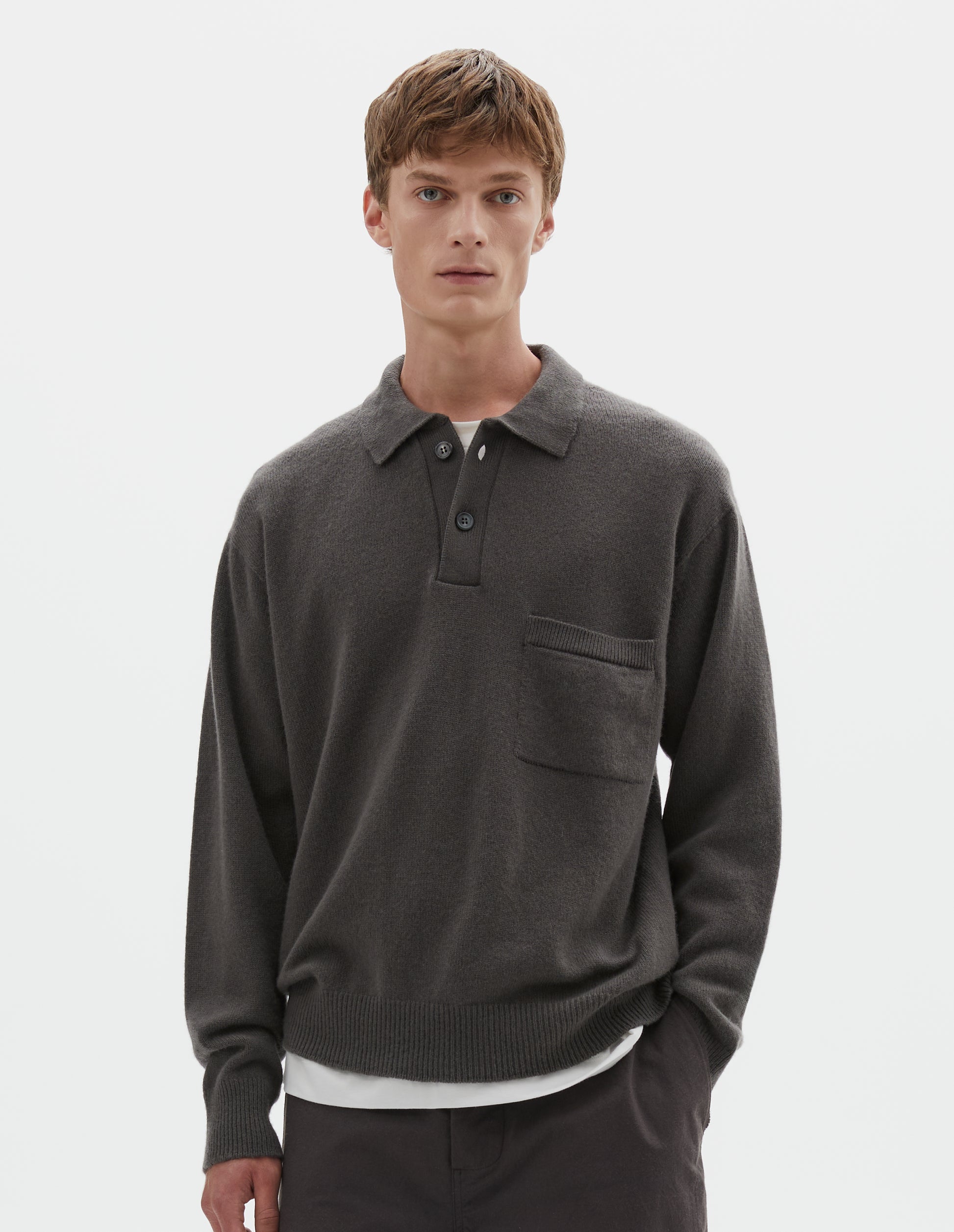 Cropped body image, front view of model wearing dark grey merino cashmere polo shirt with dropped sleeve, integral chest pocket, horn button closure and rib collar, cuffs and hem. Styled over white t-shirt, with brown shorts.