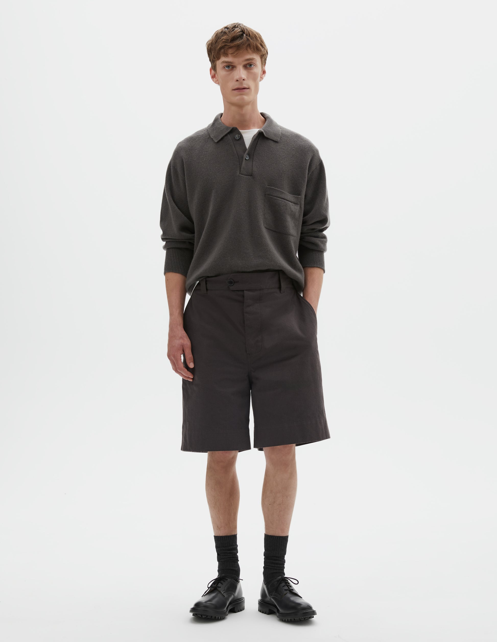 Full body image, front view model wearing dark brown cotton shorts with belt loops, button fly with button tab fastening and slant pockets. Styled with grey collared jumper tucked into waistband and styled over a white t shirt, with black socks and black leather shoes. 