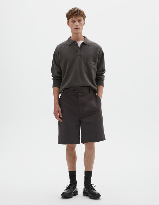 Full body image, front view model wearing dark brown cotton shorts with belt loops, button fly with button tab fastening and slant pockets. Styled with grey collared jumper tucked into waistband and styled over a white t shirt, with black socks and black leather shoes. 