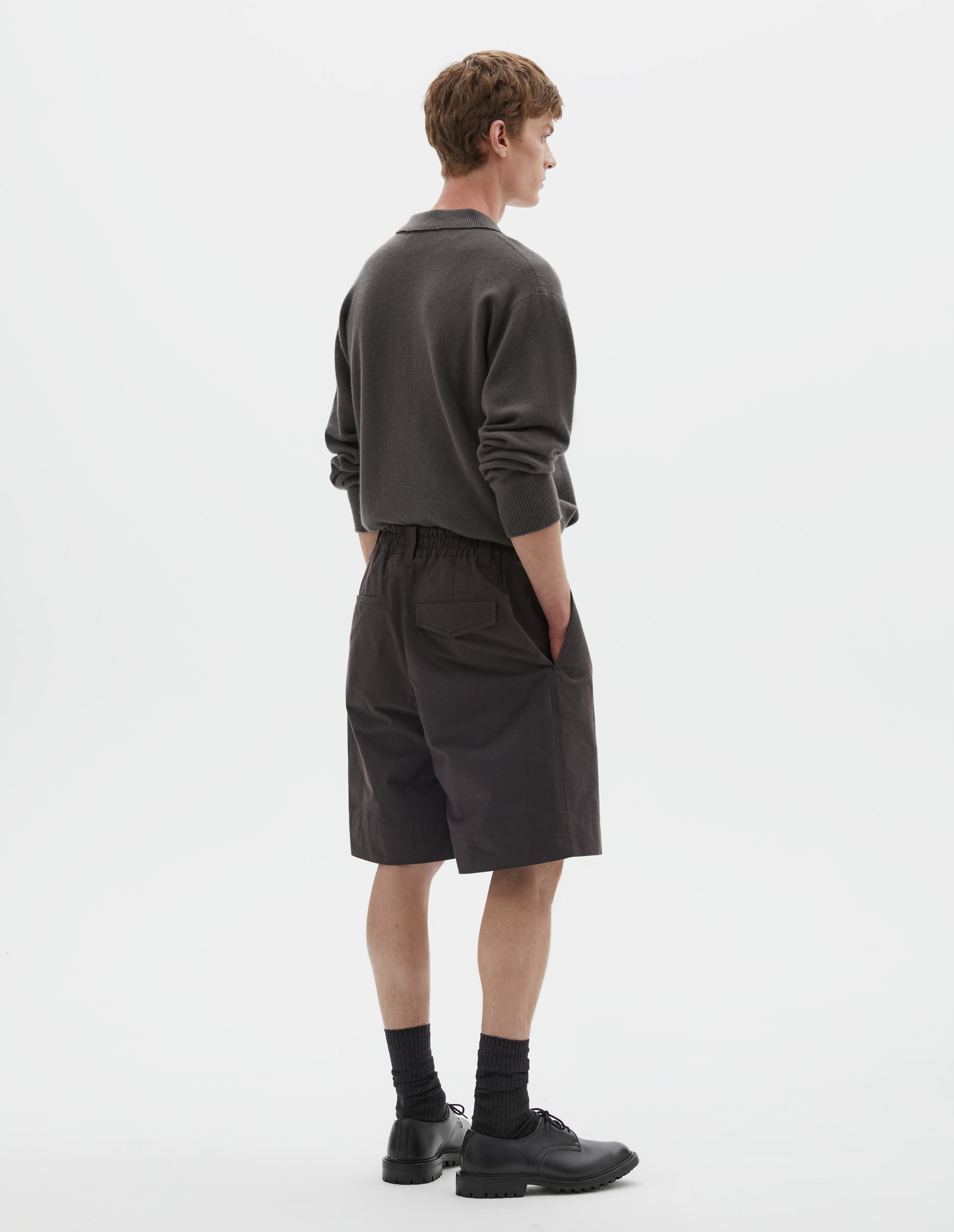 Full body image, back view of model wearing dark brown relaxed shorts with half elasticated waistband, belt loops, slant side pockets and concealed button fastening flap back pocket. Styled with collared grey jumper tucked into waistband, with black socks and black leather shoes.