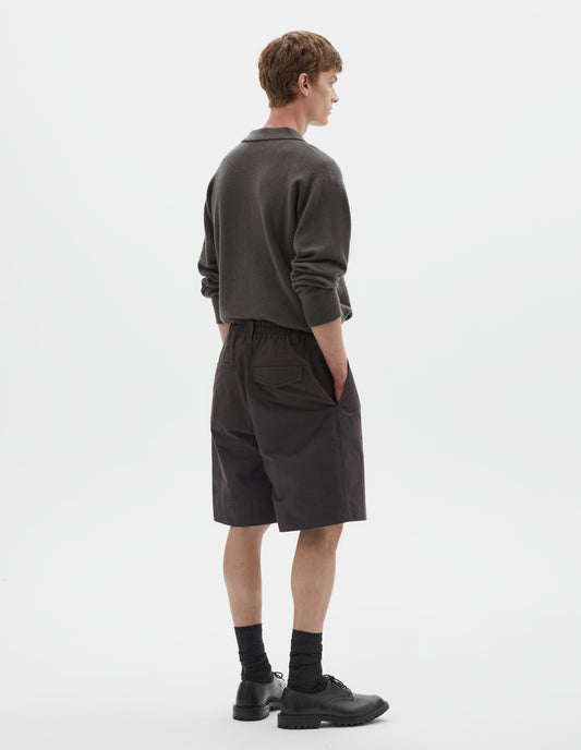 Full body image, back view of model wearing dark brown relaxed shorts with half elasticated waistband, belt loops, slant side pockets and concealed button fastening flap back pocket. Styled with collared grey jumper tucked into waistband, with black socks and black leather shoes.