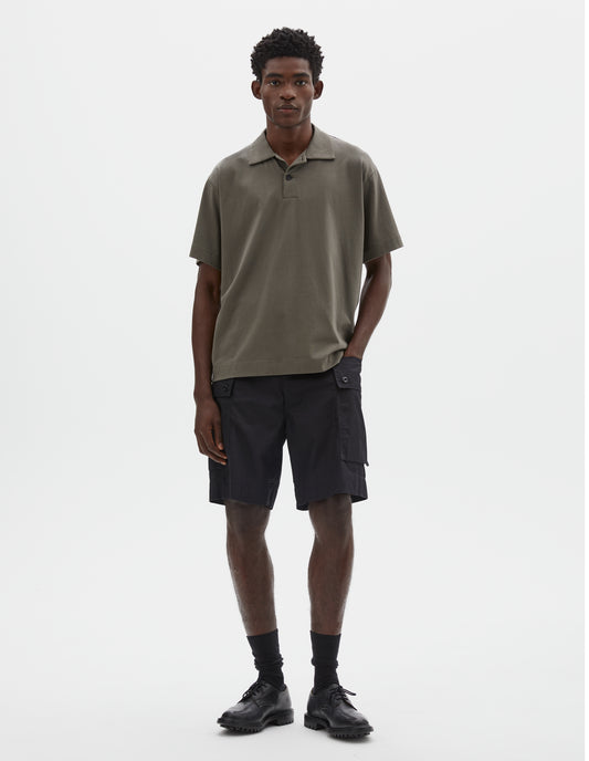 Full body image, front view of model wearing greyish green cotton short sleeve  polo shirt with wide placket and horn buttons. Styled with black cargo shorts, black socks and black leather shoes.