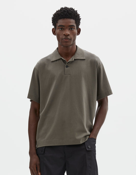 Cropped image, front view of model wearing greyish green cotton short sleeve polo shirt with wide placket and horn buttons. Styled with black cargo shorts.