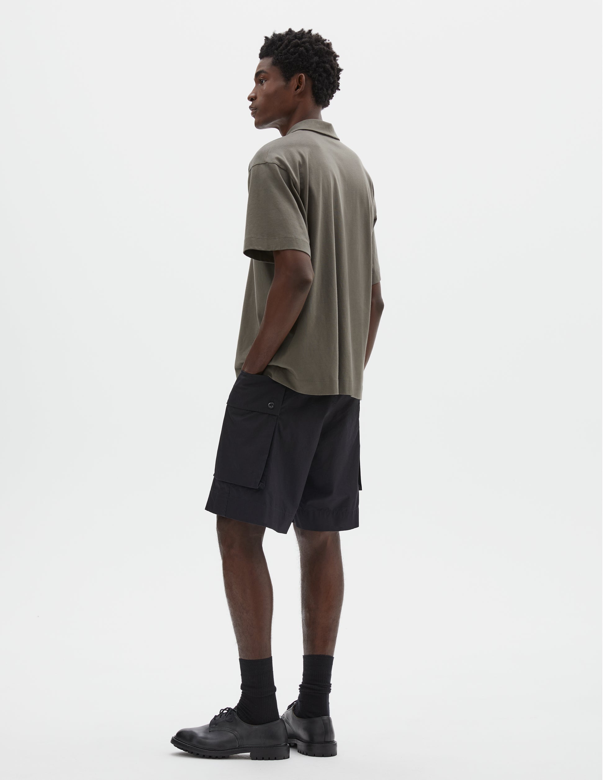 Full body image, back view of model wearing short sleeve greyish green cotton polo shirt. Styled with black cargo shorts, black socks and black leather shoes.  