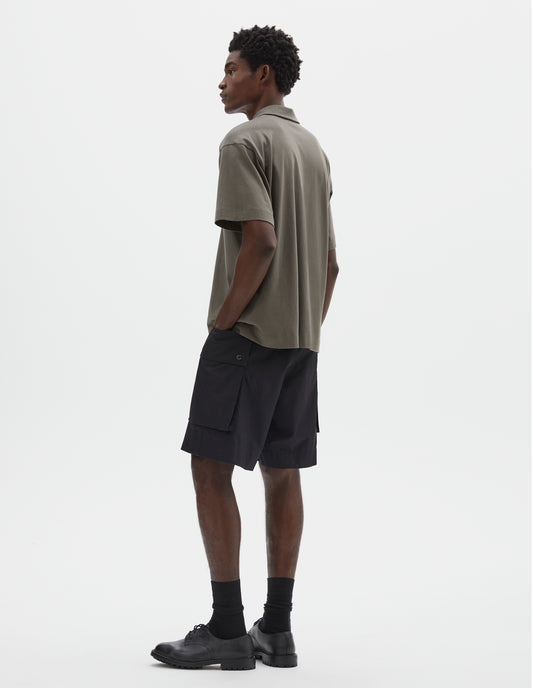Full body image, back view of model wearing short sleeve greyish green cotton polo shirt. Styled with black cargo shorts, black socks and black leather shoes.  