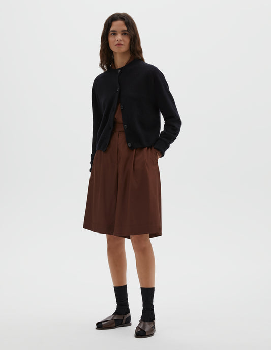 Full body image, front view of model wearing dark navy long sleeve merino cashmere cardigan with contrast rib neckline, cuffs and hem and dark horn buttons. Styled with brown shirt, matching brown pleated shorts, black socks and brown leather sandals. 