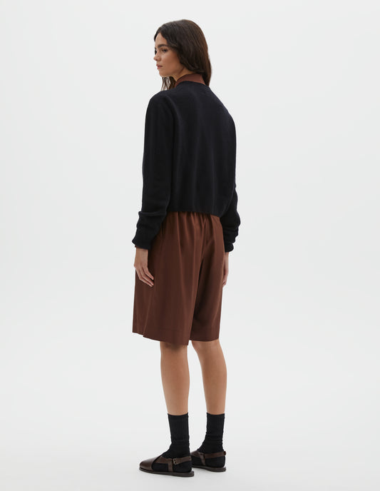 Full body image, back view of model wearing dark navy long sleeve merino cashmere cardigan with contrast rib neckline, cuffs and hem. Styled with brown pleated shorts, black socks and brown leather sandals. 