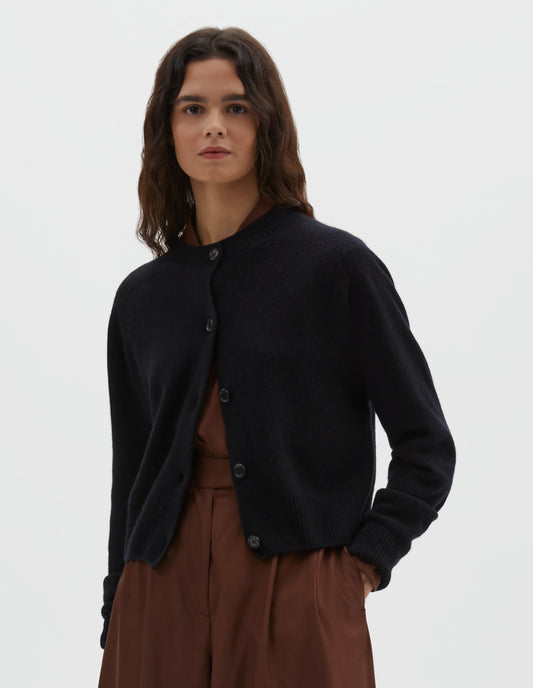 Cropped body image, front view of model wearing dark navy long sleeve merino cashmere cardigan with contrast rib neckline, cuffs and hem and dark horn buttons. Styled with brown shirt and matching brown pleated shorts.