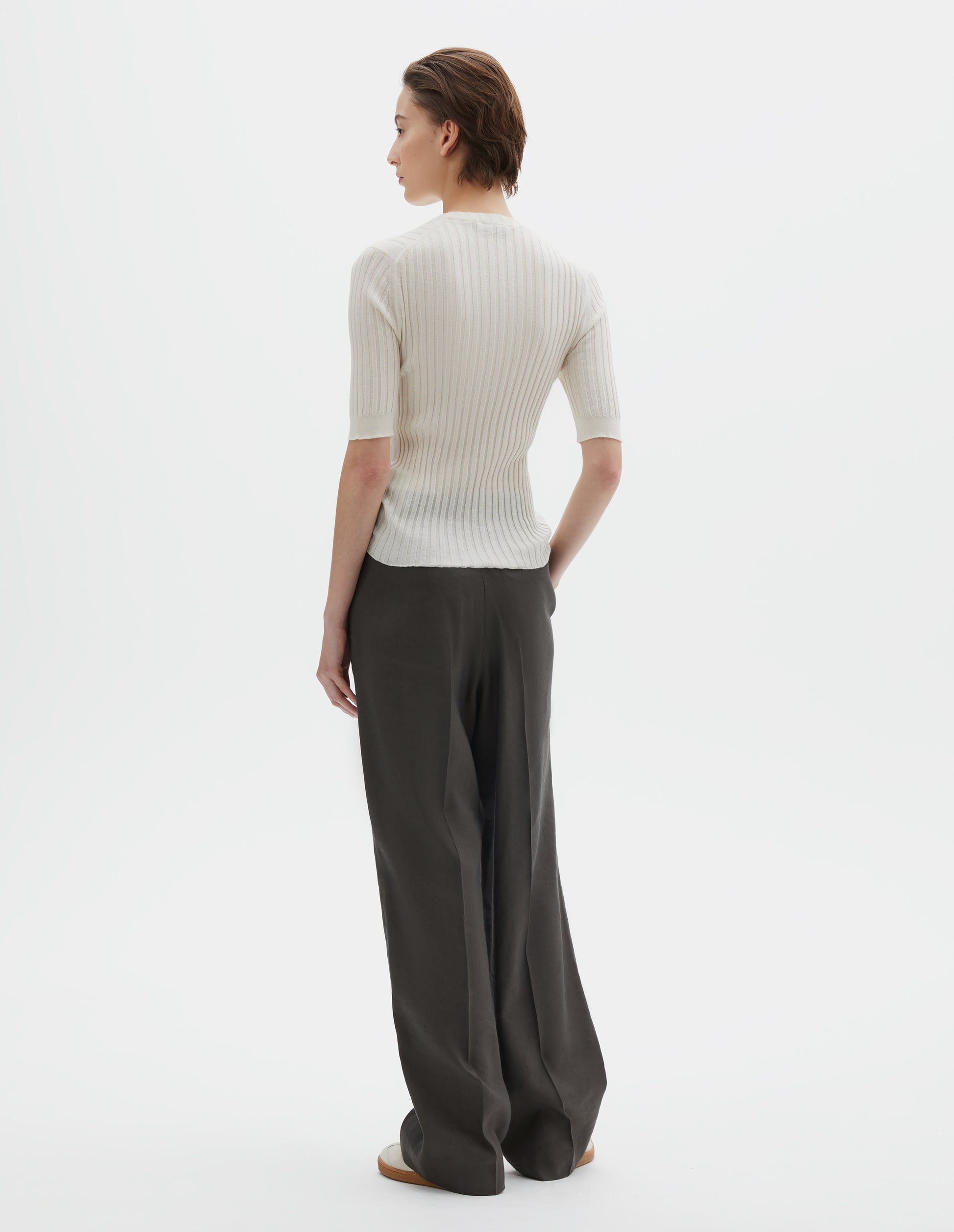Full body image, back view of model wearing beige knitted ribbed t-shirt with round neck and fine rib sleeve cuffs. Styled with wide greyish-green  trousers and beige nubuck trainers.