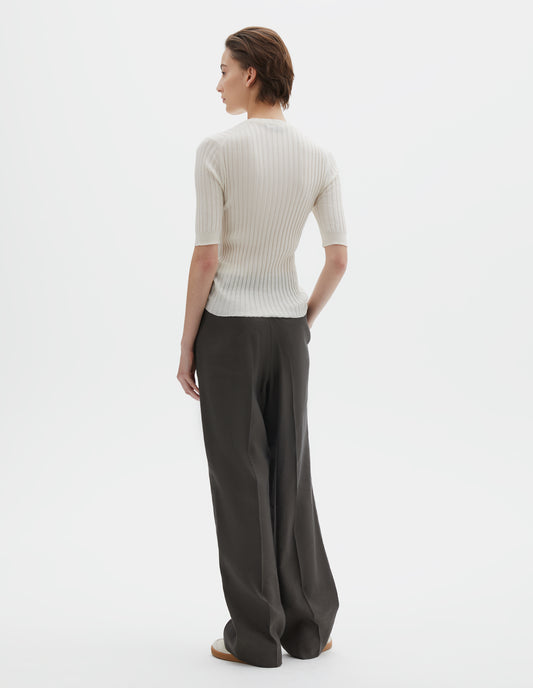 Full body image, back view of model wearing beige knitted ribbed t-shirt with round neck and fine rib sleeve cuffs. Styled with wide greyish-green  trousers and beige nubuck trainers.