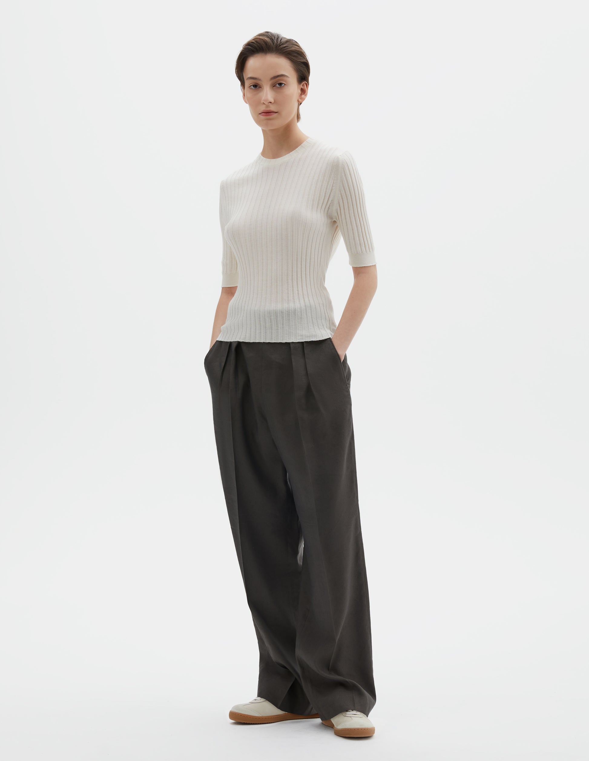 Full body image, front view of model wearing beige knitted ribbed t-shirt with round neck and fine rib sleeve cuffs. Styled with wide greyish-green  trousers and beige nubuck trainers.