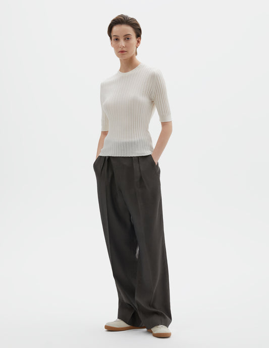 Full body image, front view of model wearing beige knitted ribbed t-shirt with round neck and fine rib sleeve cuffs. Styled with wide greyish-green  trousers and beige nubuck trainers.