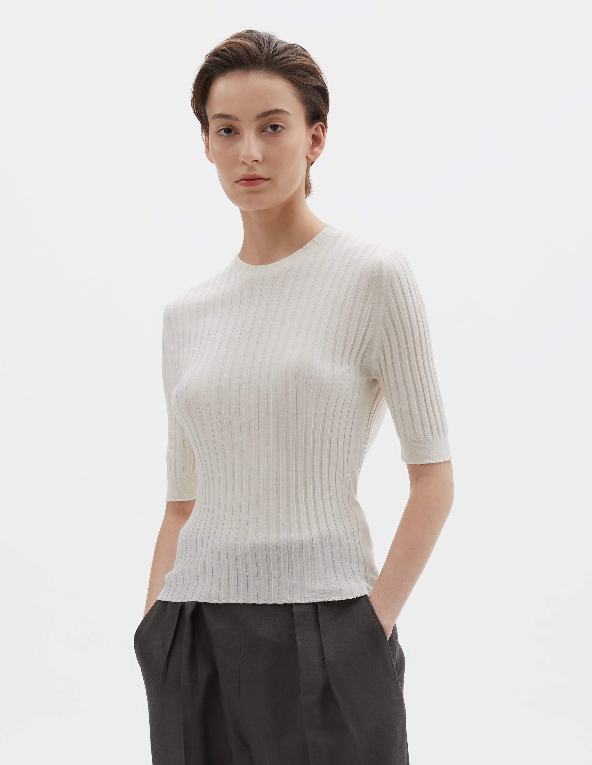 Cropped body image, front view of model wearing beige knitted ribbed t-shirt with round neck and fine rib sleeve cuffs. Styled with greyish-green trousers.
