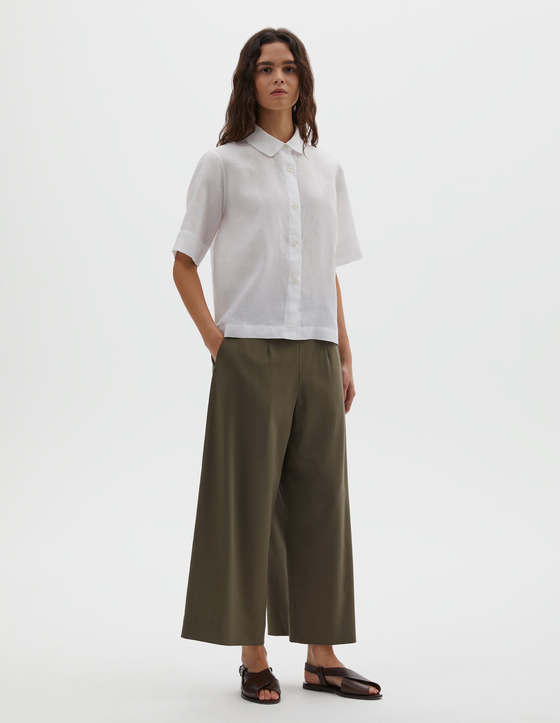 Full body image, front view of model wearing  wearing white linen short sleeve shirt, with rounded collar, mother of pearl buttons and side split hem. Styled with brown culottes and brown leather sandals.