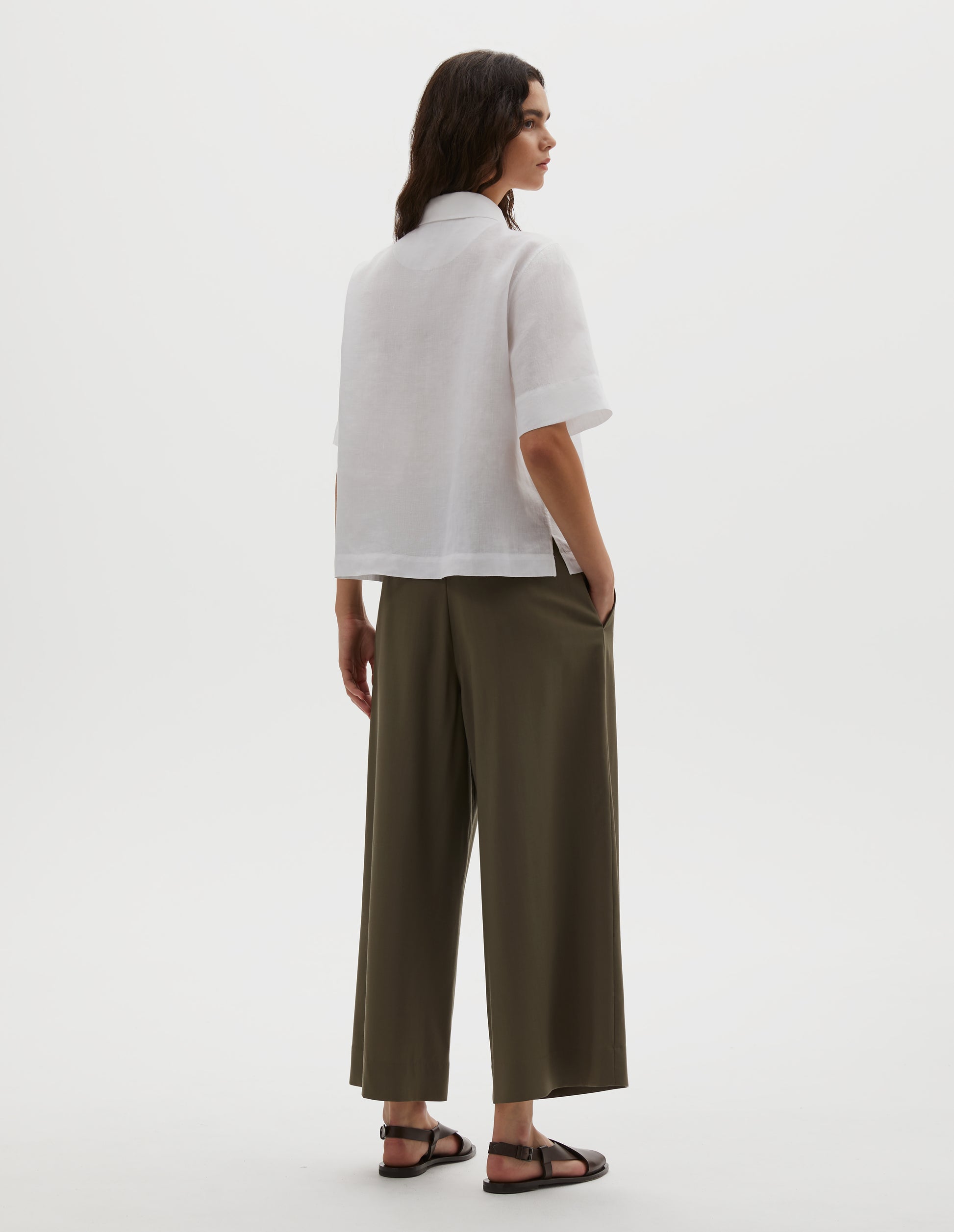 Full body image, back view of model wearing white linen short sleeve shirt, with side split hem. Styled with brown culottes and brown leather sandals.