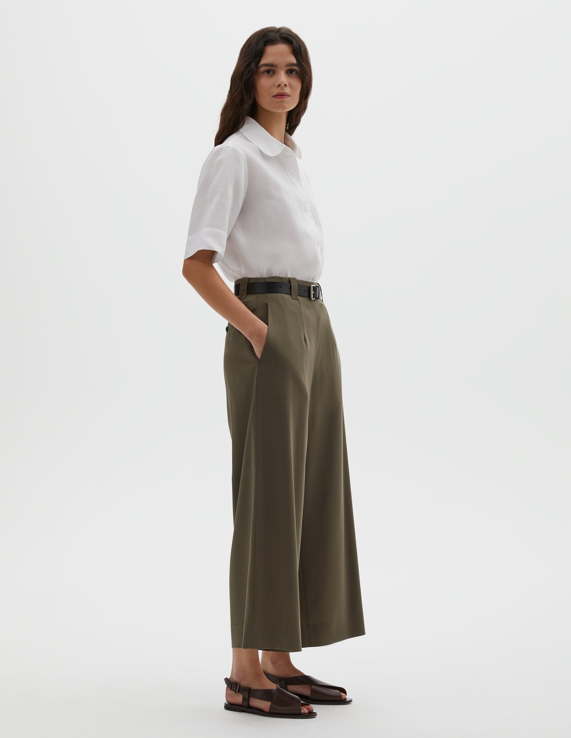 Full body image, front view of model wearing brown wool wide leg cropped culottes with concealed side zip and slant pockets. Styled with white shirt, dark brown belt and dark brown leather sandals.