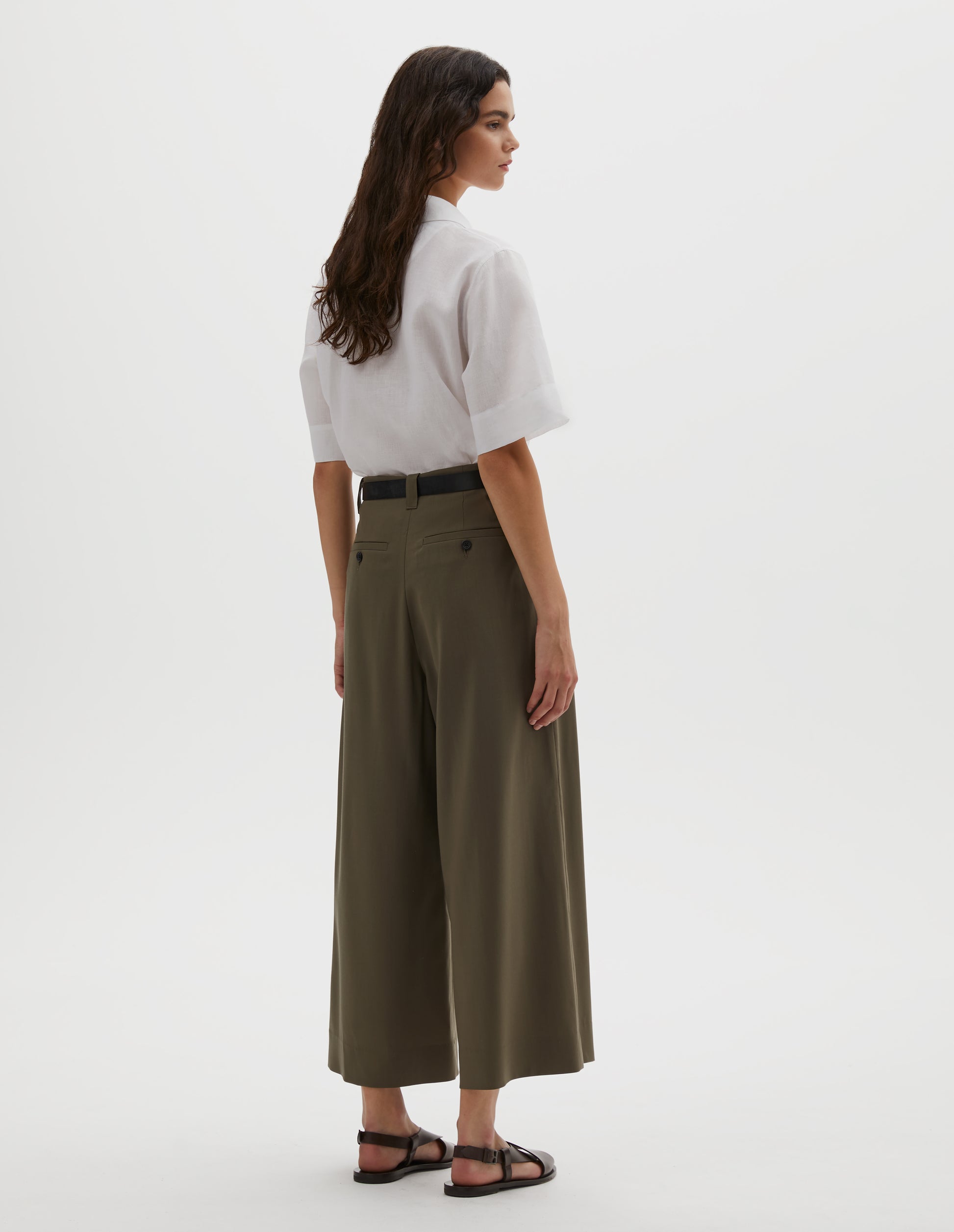 Full body image, back view of model wearing brown wool wide leg cropped culottes with concealed side zip, slant pockets and button-through welt back pocket. Styled with white shirt, dark brown belt and dark brown leather sandals.