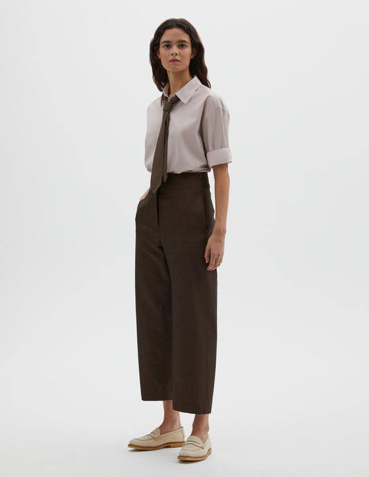 Full body image, front view of model wearing brown cotton linen trousers, with deep waistband, double horn button closure and slant side pockets. Styled with pink shirt, brown tie and beige suede loafers.