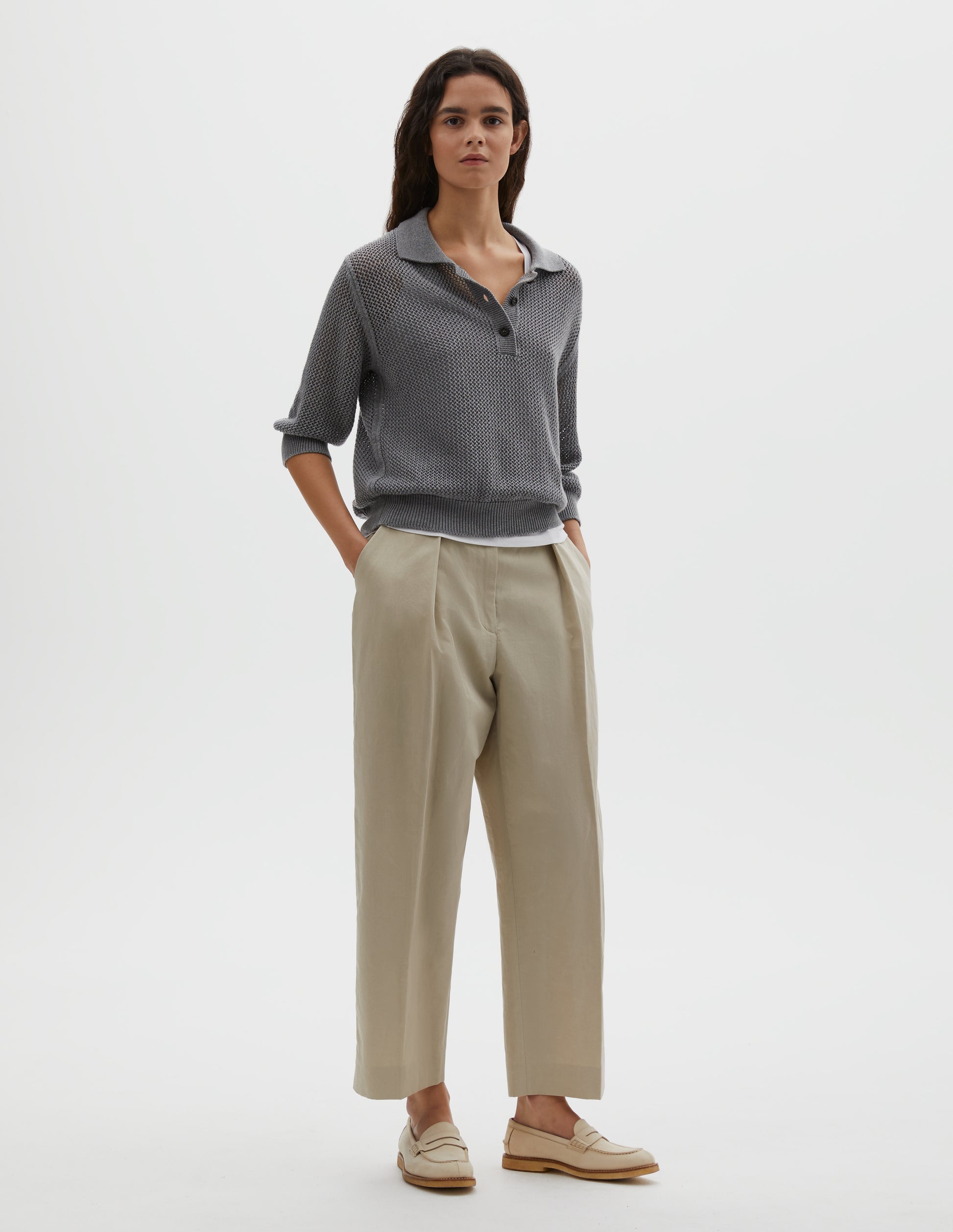 Full body image, front view of model wearing dark grey cotton cashmere pointelle polo shirt, with solid seam detailing, rib collar, placket, cuffs and hem and horn button closure. Styled over white cotton vest, with beige pleated trousers and beige suede loafers.