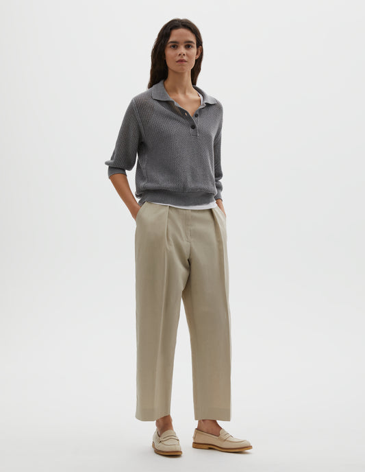 Full body image, front view of model wearing dark grey cotton cashmere pointelle polo shirt, with solid seam detailing, rib collar, placket, cuffs and hem and horn button closure. Styled over white cotton vest, with beige pleated trousers and beige suede loafers.