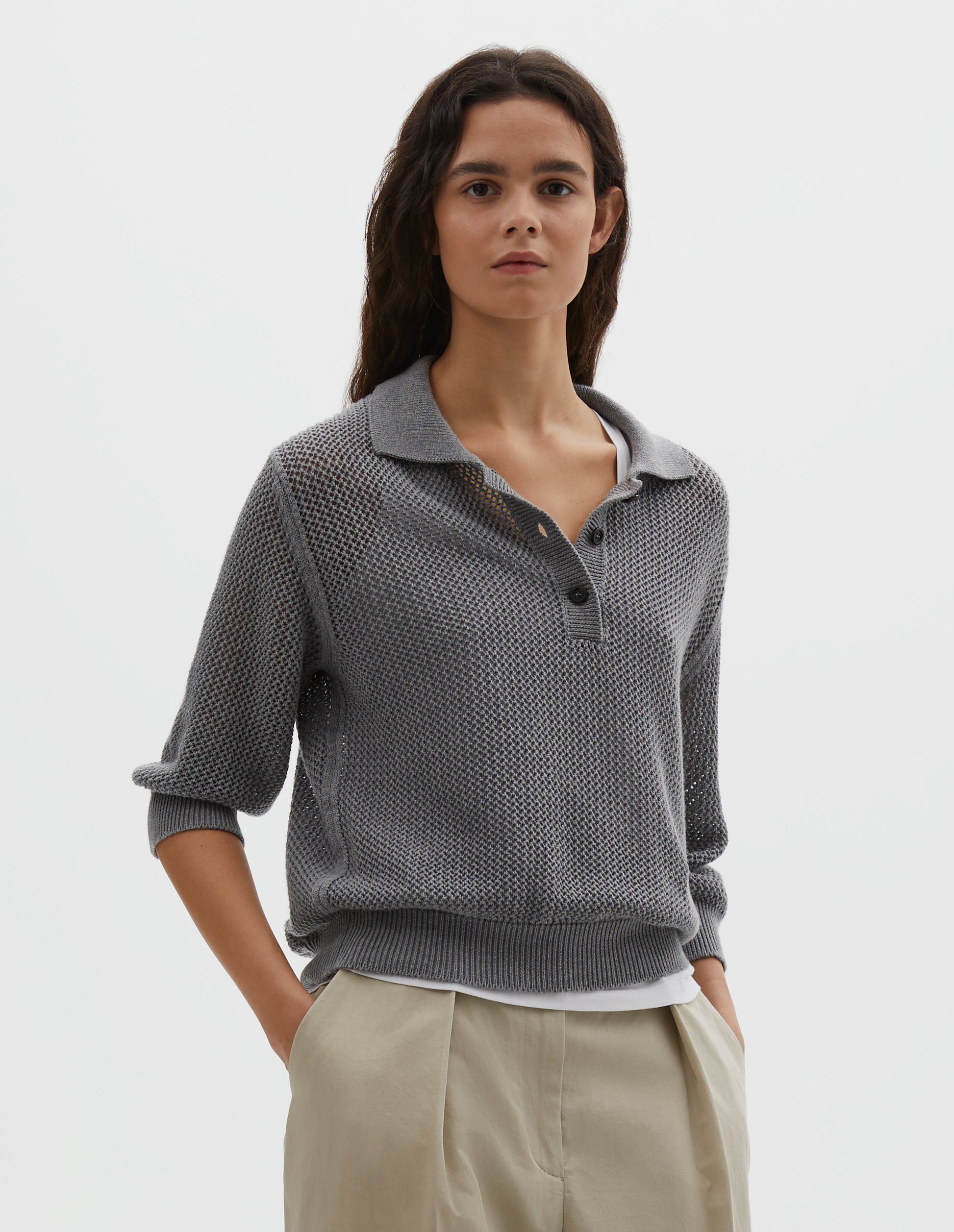 Cropped body image, front view of model wearing dark grey cotton cashmere pointelle polo shirt, with solid seam detailing, rib collar, placket, cuffs and hem and horn button closure. Styled over white cotton vest, with beige pleated trousers.