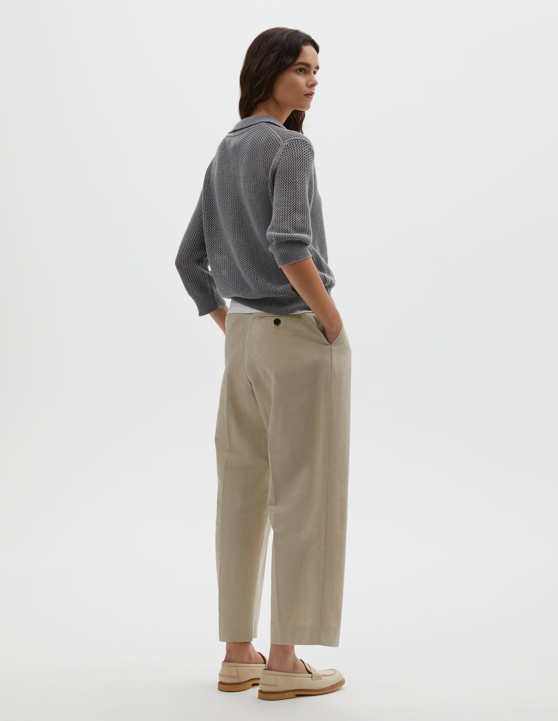 Full body image, back view of model wearing dark grey cotton cashmere pointelle polo shirt, with solid seam detailing, rib collar, placket, cuffs and hem. Styled over white cotton vest, with beige pleated trousers and beige suede loafers.