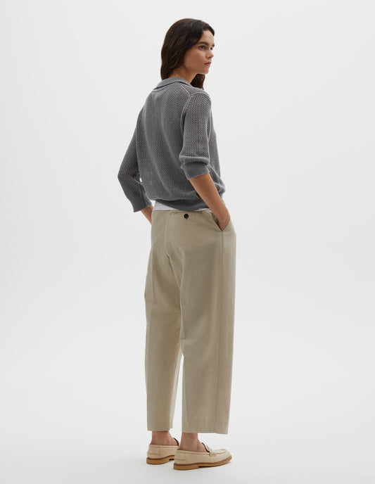 Full body image, back view of model wearing dark grey cotton cashmere pointelle polo shirt, with solid seam detailing, rib collar, placket, cuffs and hem. Styled over white cotton vest, with beige pleated trousers and beige suede loafers.