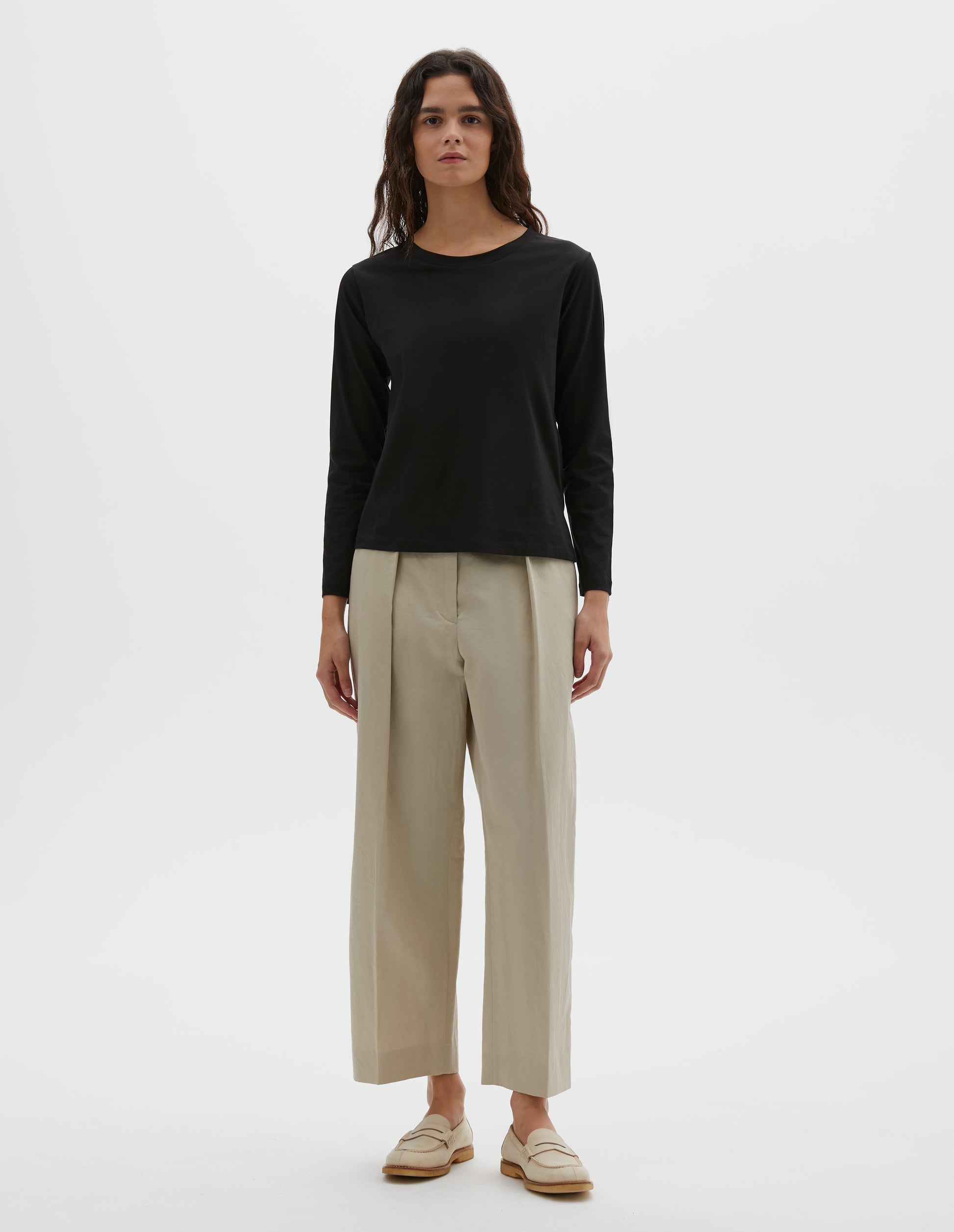 Full body image, front view of model wearing black cotton wide neck long sleeve with boxy cut. Styled with beige pleated trousers and beige suede loafers.