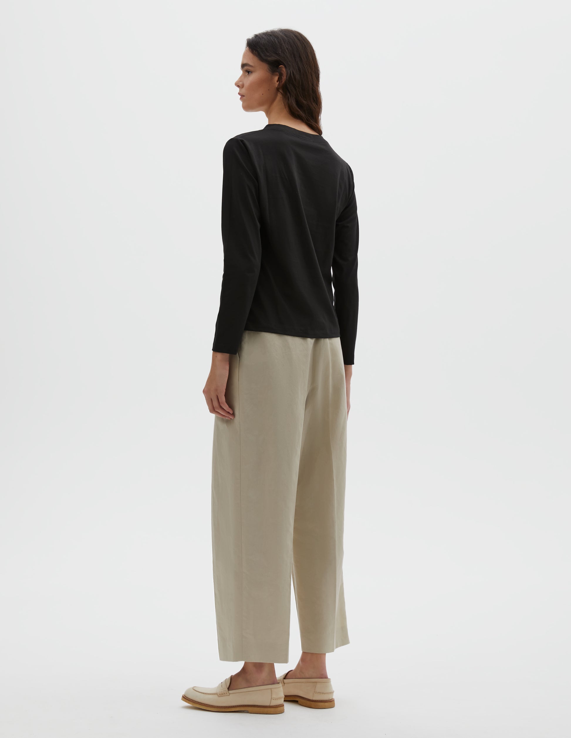 Full body image, back view of model wearing black cotton wide neck long sleeve with boxy cut. Styled with beige pleated trousers and beige suede loafers.