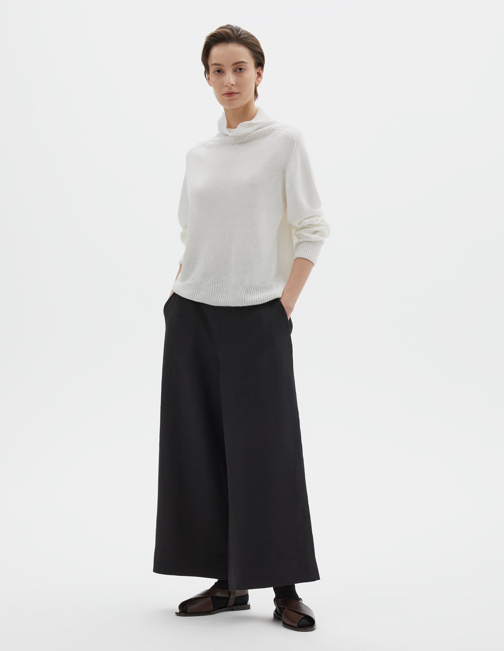 Full body image, front view of model wearing off white linen cotton roll neck jumper with long saddle sleeves and contrast rib on neck, cuffs and hem. Styled with black culotte trousers, black socks and brown leather sandals.