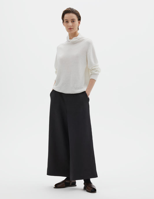 Full body image, front view of model wearing black wide leg cropped culottes with concealed side zip and slant pockets. Styled with white roll neck jumper, black socks and dark brown leather sandals.