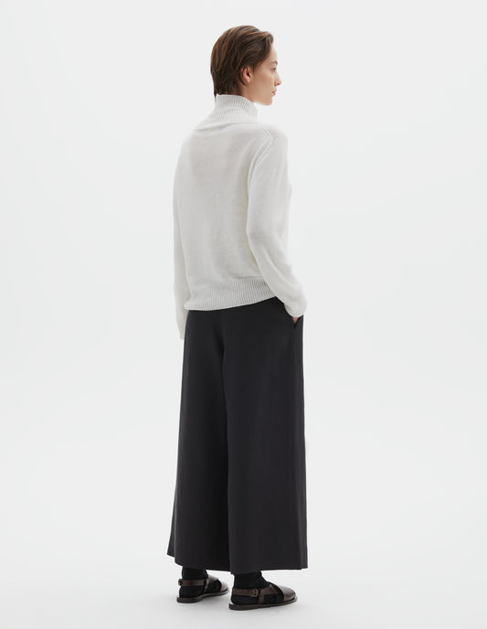 Full body image, back view of model wearing black wide leg cropped culottes with concealed side zip, slant pockets and button-through welt back pocket. Styled with white roll neck jumper, black socks and dark brown leather sandals.