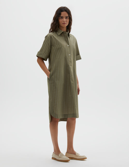 Full body image, front view of model wearing green with light blue pin stripe cotton silk shirt dress, with raised stitch placket, concealed side pockets, two patch chest pockets and mother of pearl button closure. Styled with beige shoes. 
