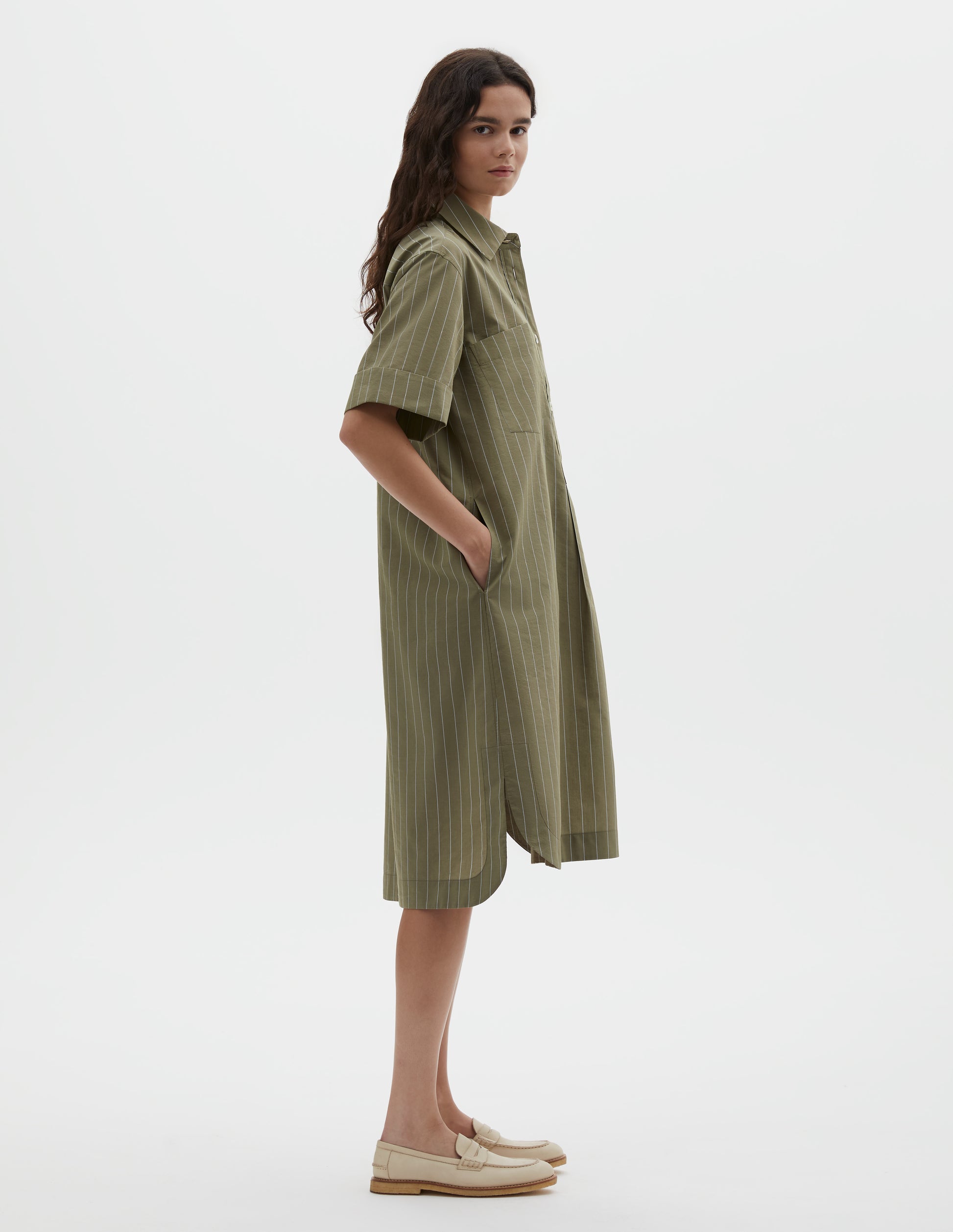 Full body image, side view of model wearing green with light blue pin stripe cotton silk shirt dress, with raised stitch placket, concealed side pockets, two patch chest pockets and mother of pearl button closure. Styled with beige shoes. 