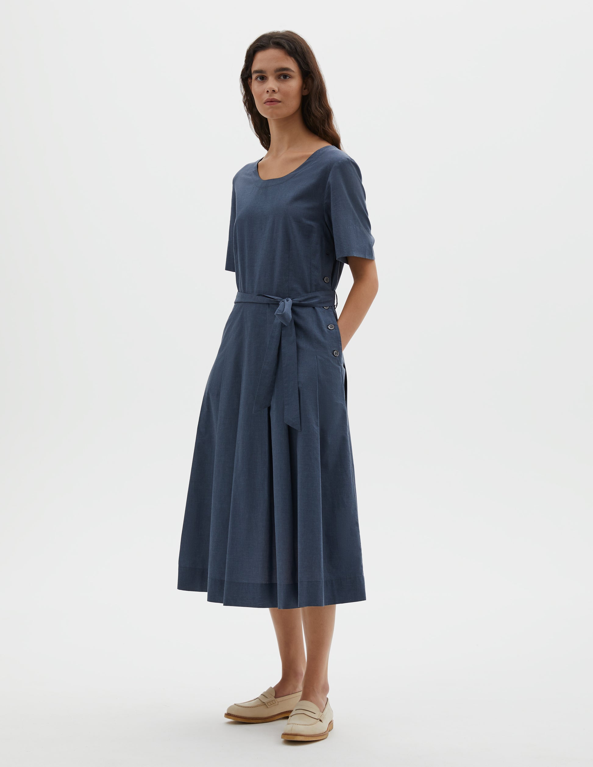 Full body image, front view of model wearing blue cotton chambray dress, with t-shirt sleeves, deep hem, stitched down pleats, self tie, side pockets and mother of pearl button detailing on one side. Styled with beige suede loafers.