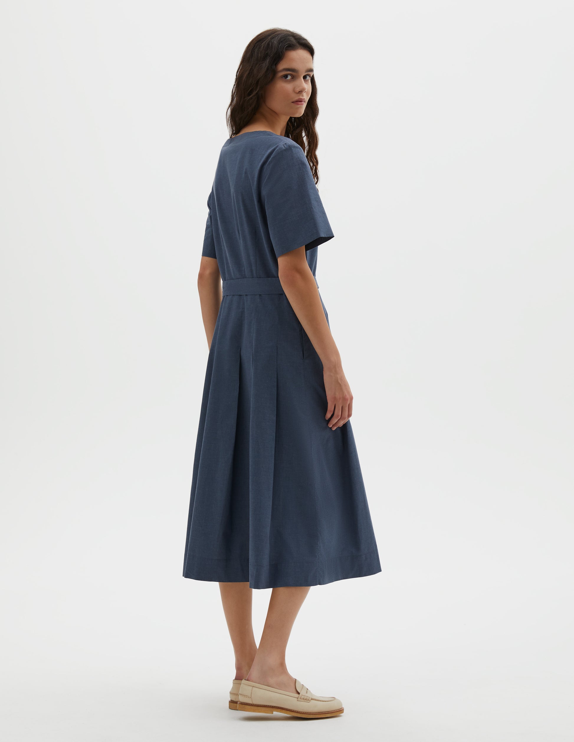 Full body image, back view of model wearing blue cotton chambray dress, with t-shirt sleeves, deep hem, stitched down pleats, self tie and side pockets. Styled with beige suede loafers.