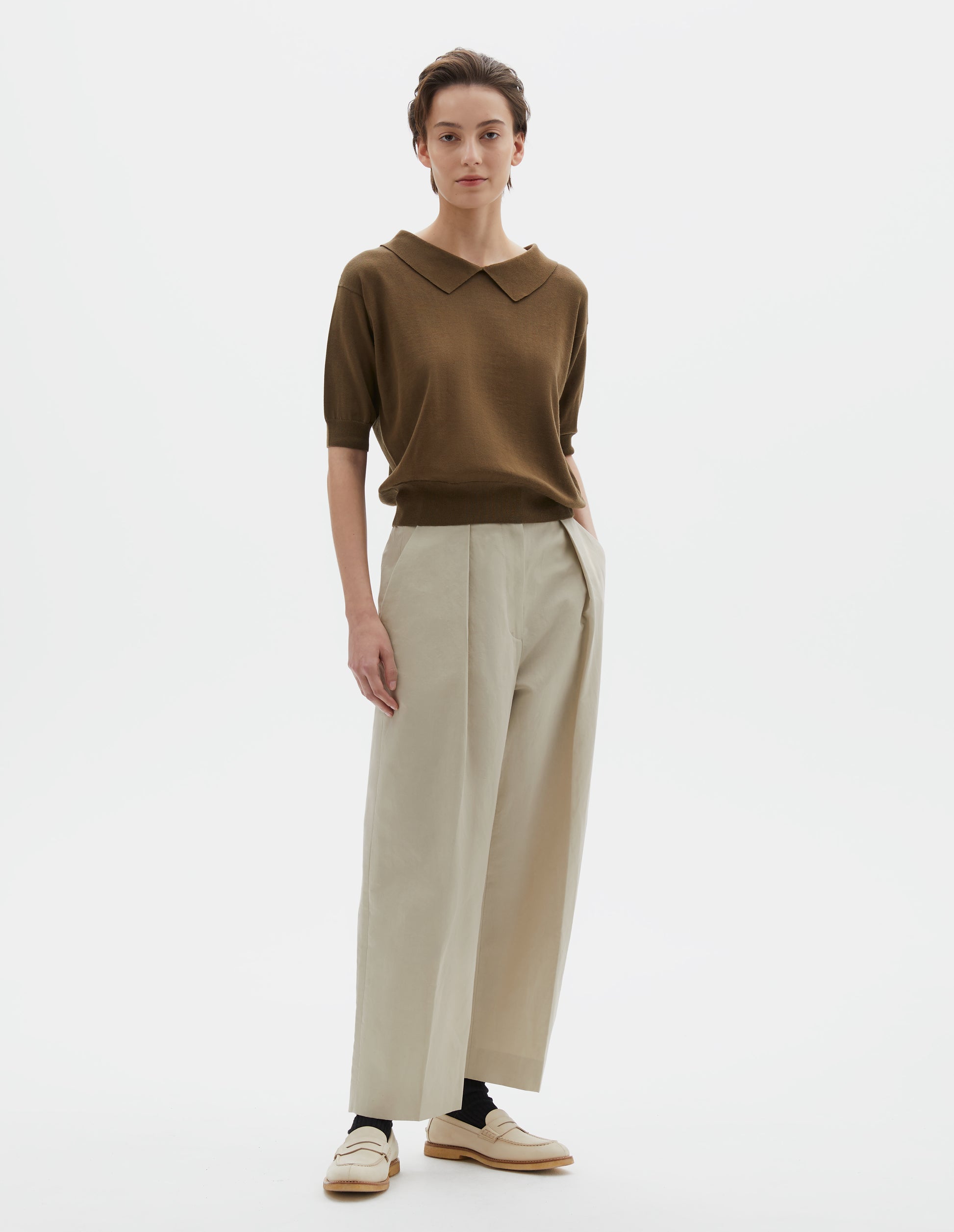 Full body image, front view of model wearing regular fit green split collar knitted t-shirt with ribbed cuffs and hem. Styled with beige trousers, black socks and beige leather loafers.