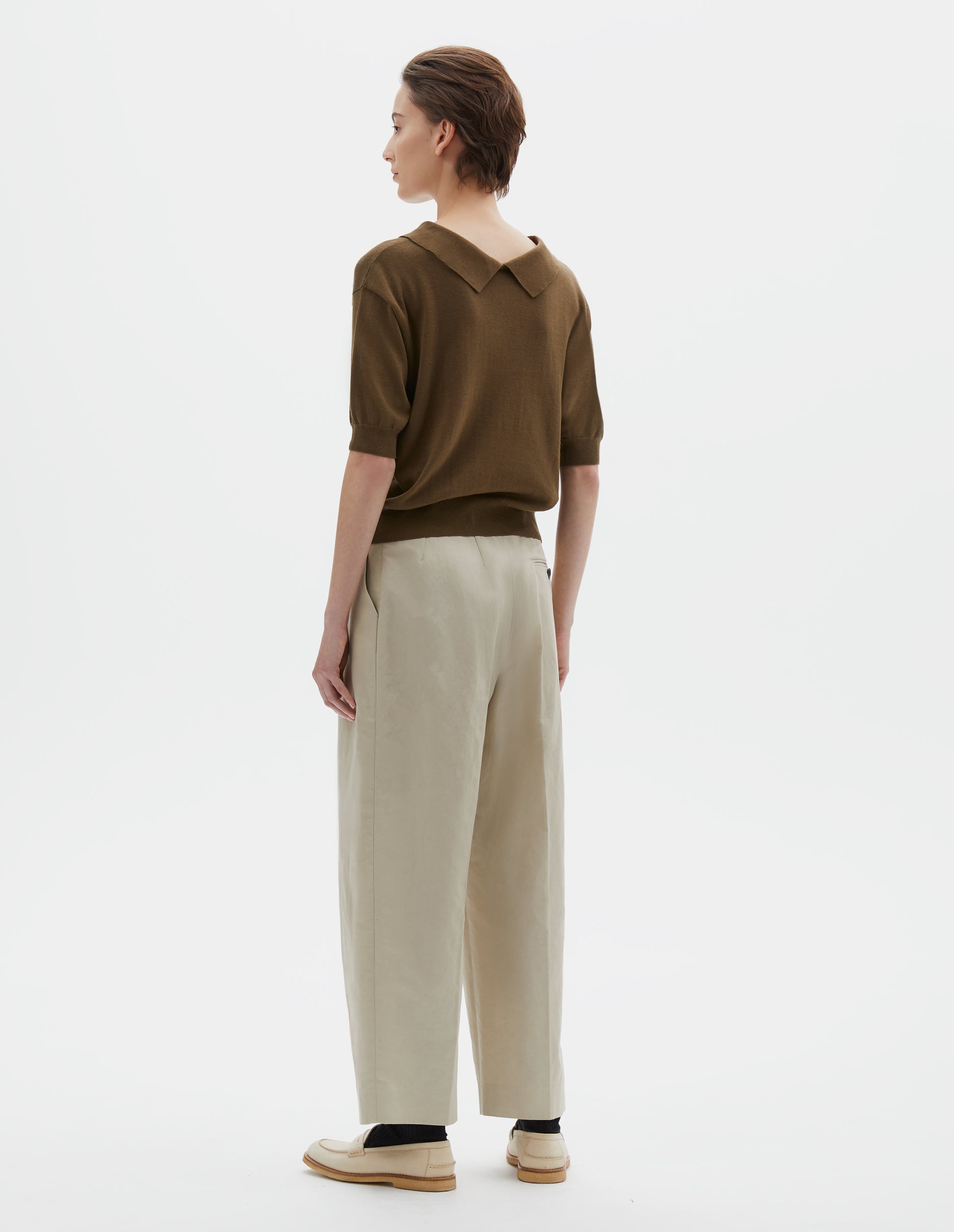 Full body image, back view of model wearing regular fit green split collar knitted t-shirt with ribbed cuffs and hem. Styled with beige trousers, black socks and beige leather loafers.