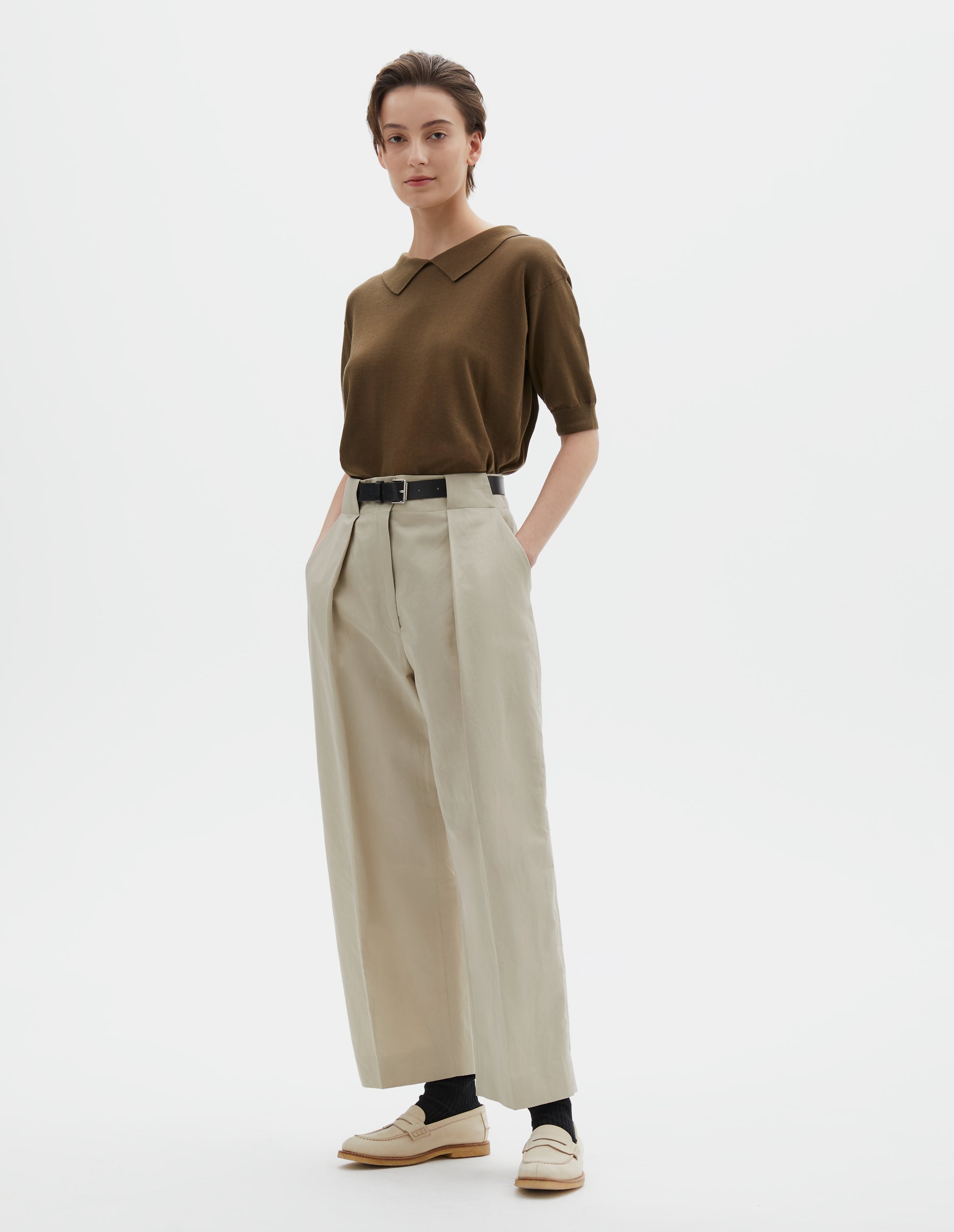 Full body image, front view of model wearing beige cotton linen blend trousers, single pleats and grown-on waistband with large belt loops and horn button. Styled with greenish brown collared knitted t-shirt , dark brown belt, black socks and beige shoes.