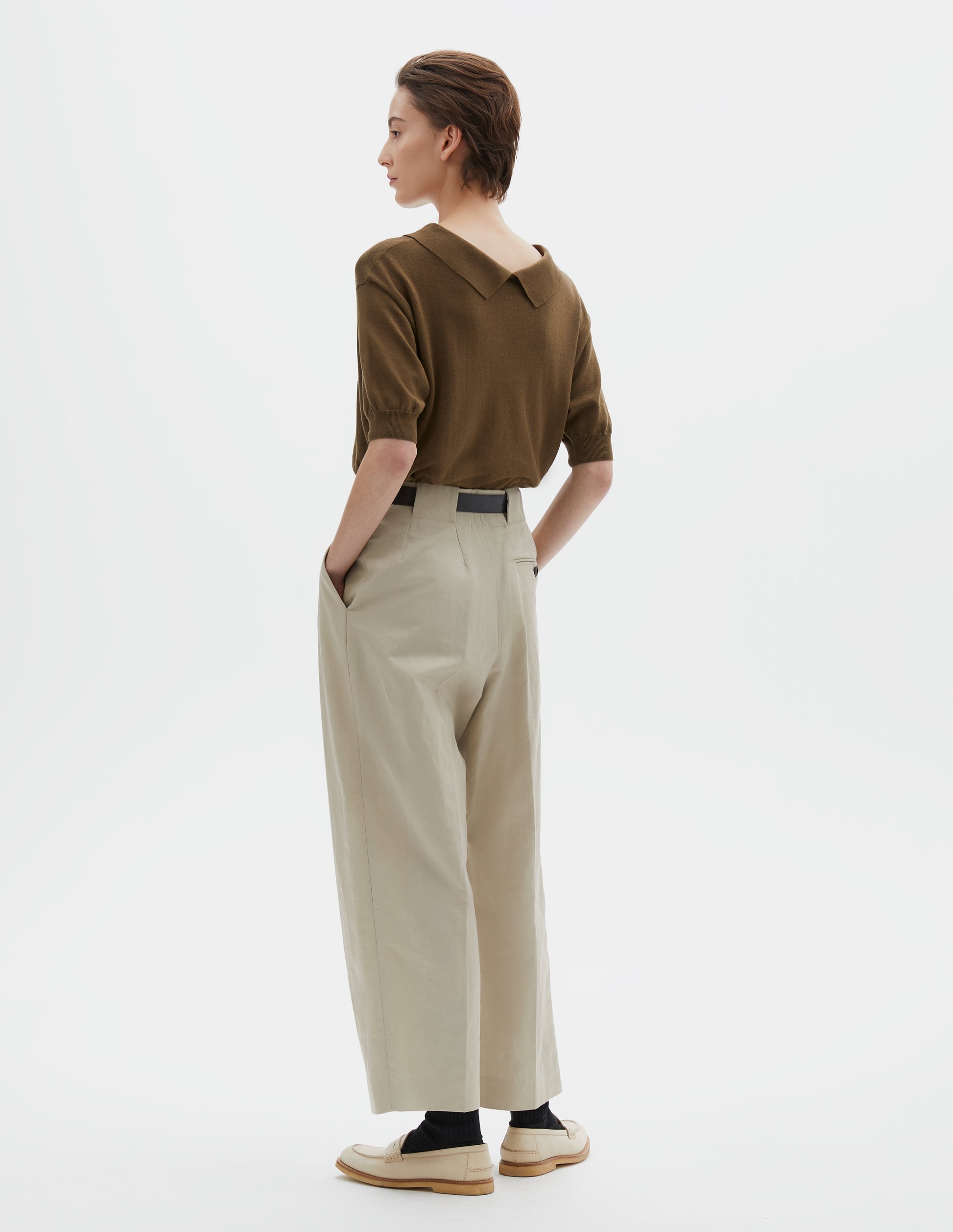 Full body image, back view of model wearing beige cotton linen blend trousers, grown-on waistband with large belt loops and a button-through jet back pocket and horn button. Styled with greenish brown collared knitted t-shirt, dark brown belt, black socks and beige shoes.