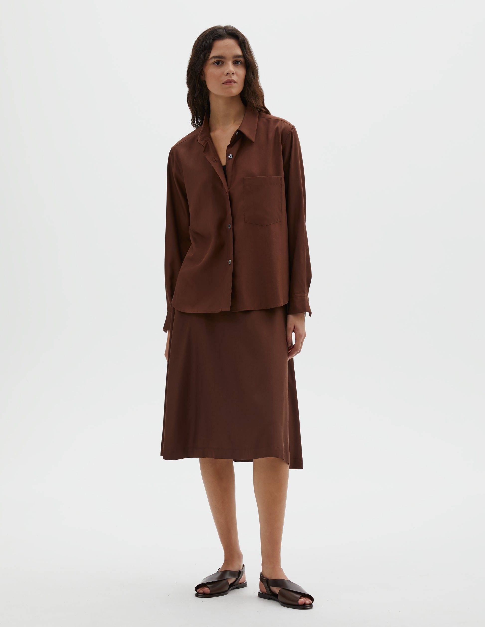 Full body image, front view of model wearing  brown shirt with raised placket, horn buttons and patch pocket on chest. Styled with matching brown skirt and dark brown leather sandals.