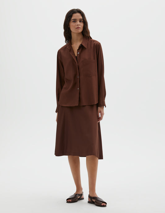 Full body image, front view of model wearing  brown shirt with raised placket, horn buttons and patch pocket on chest. Styled with matching brown skirt and dark brown leather sandals.