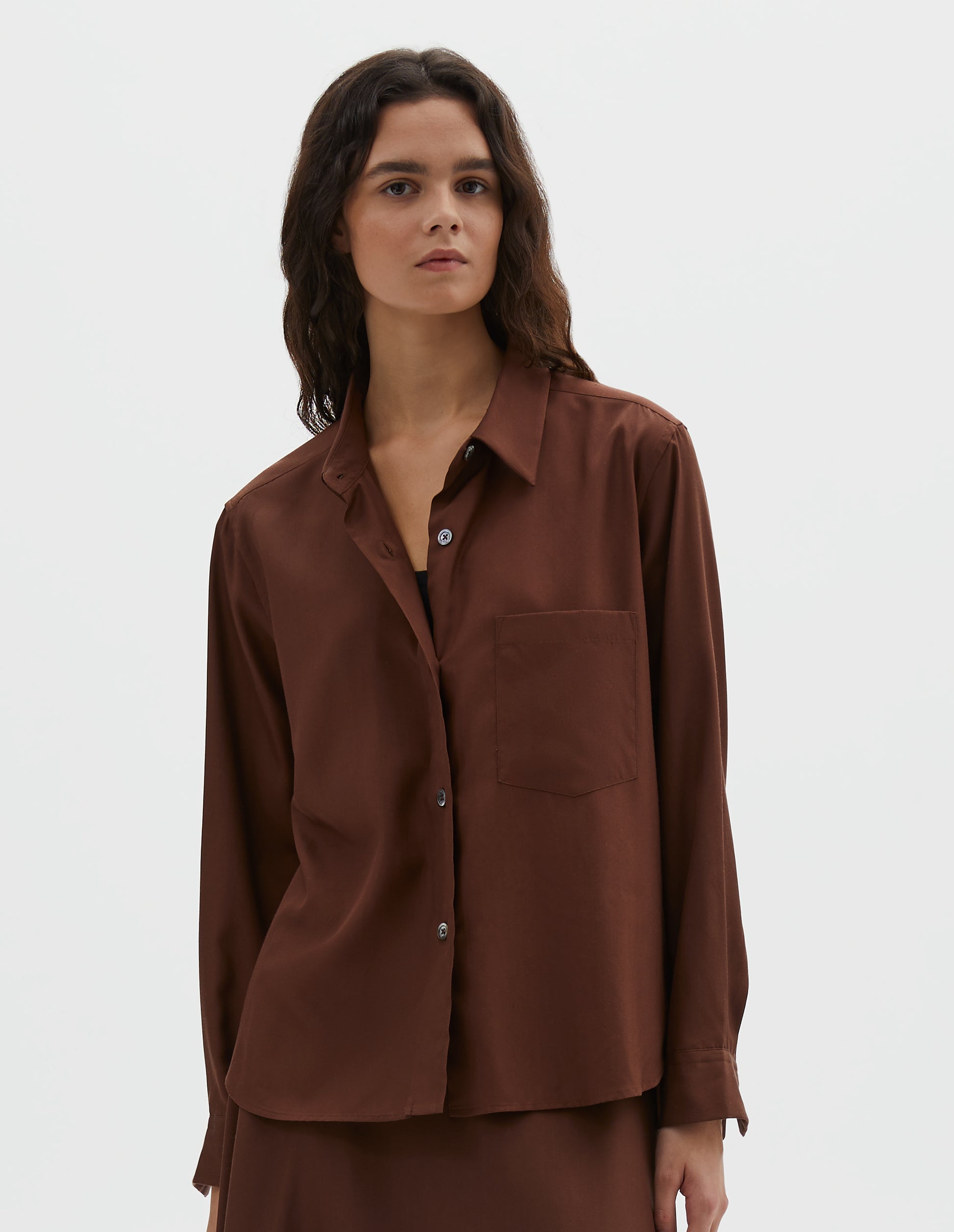 Cropped body image, front view of model wearing brown shirt with raised placket, horn buttons and patch pocket on chest. Styled with matching brown skirt.