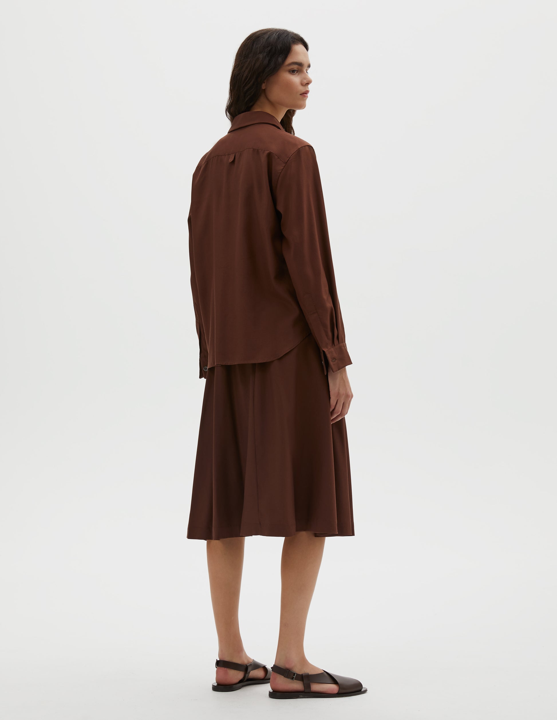 Full body image, back view of model wearing brown shirt with horn buttons on cuffs and hanger loop detail on back yoke. Styled with matching brown skirt and dark brown leather sandals.