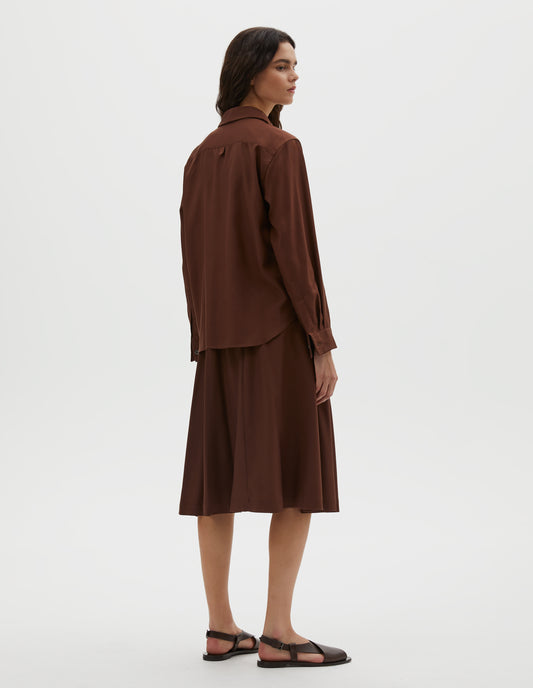 Full body image, back view of model wearing brown shirt with horn buttons on cuffs and hanger loop detail on back yoke. Styled with matching brown skirt and dark brown leather sandals.